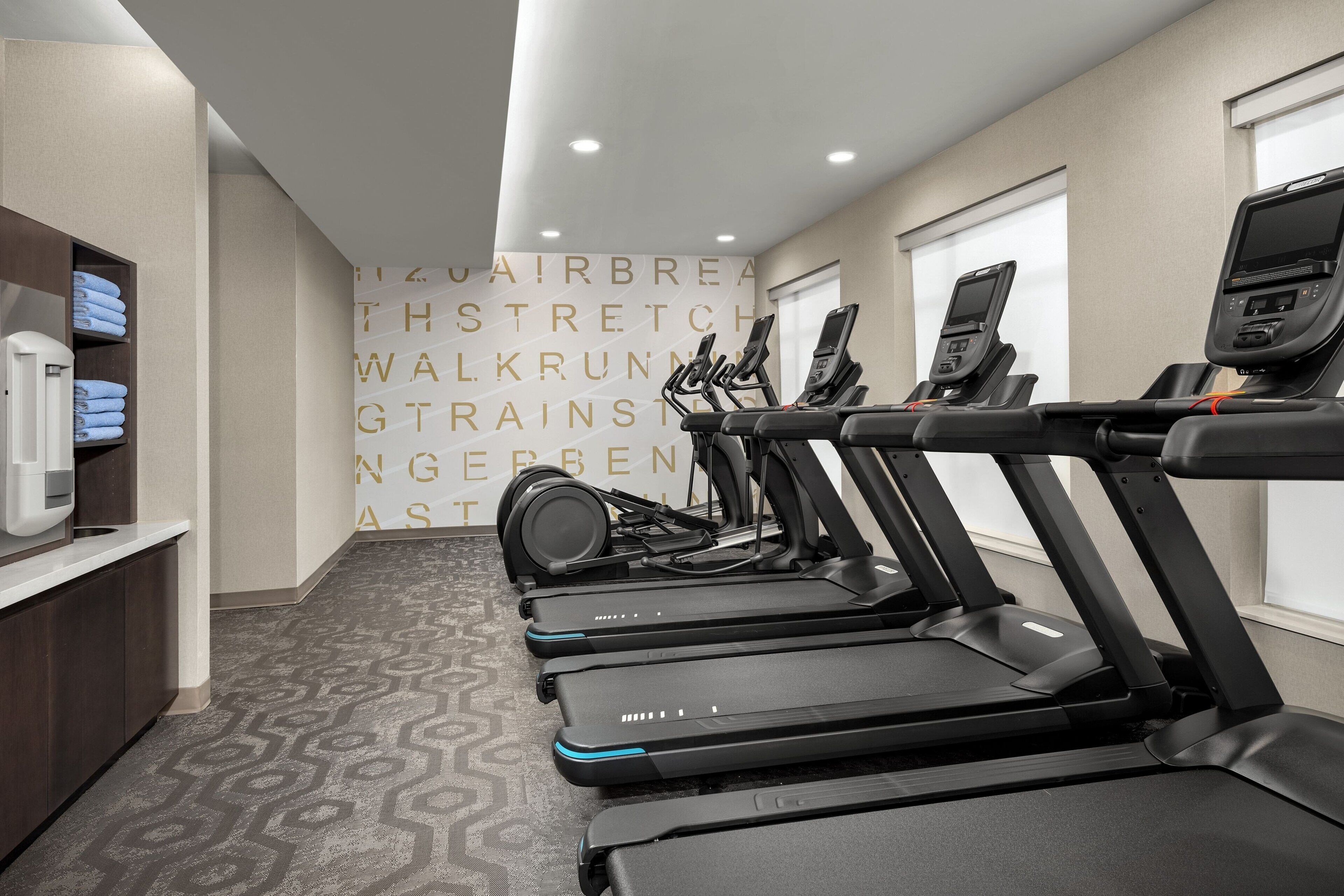 Sala de ginástica com esteiras, parede com texto motivacional, dispensador de água e carpete estampado.