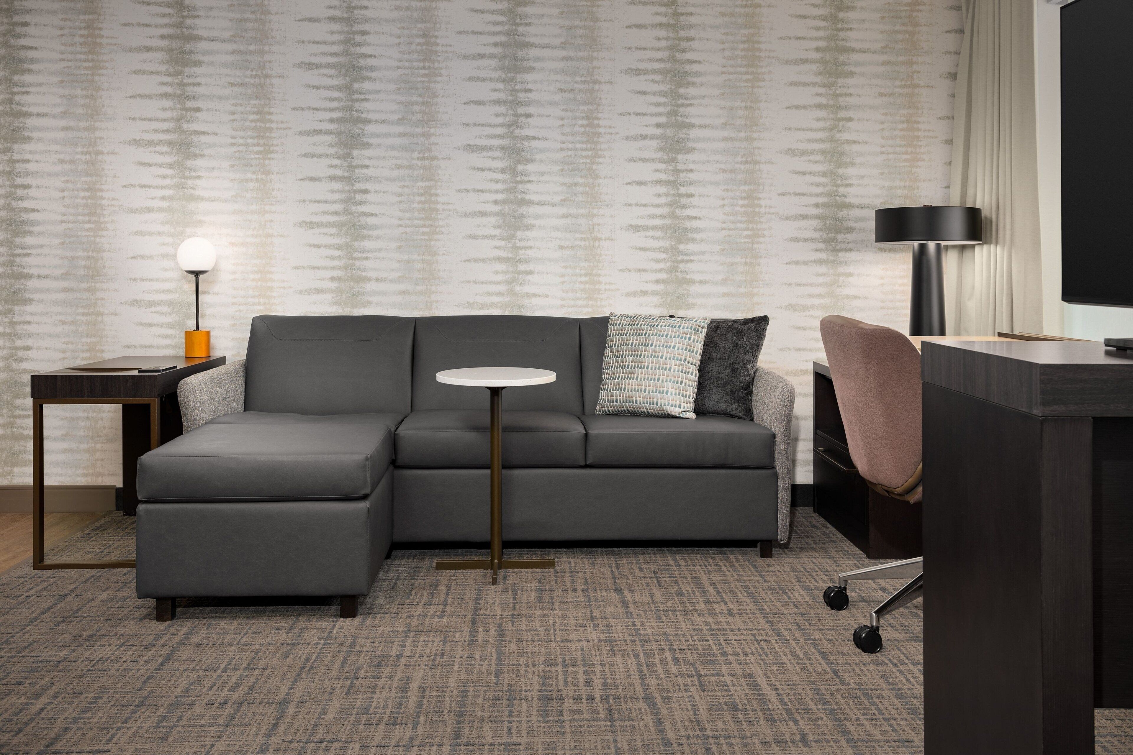 A modern living room with a grey sectional sofa, a small round table, a lamp, and a desk with a chair.