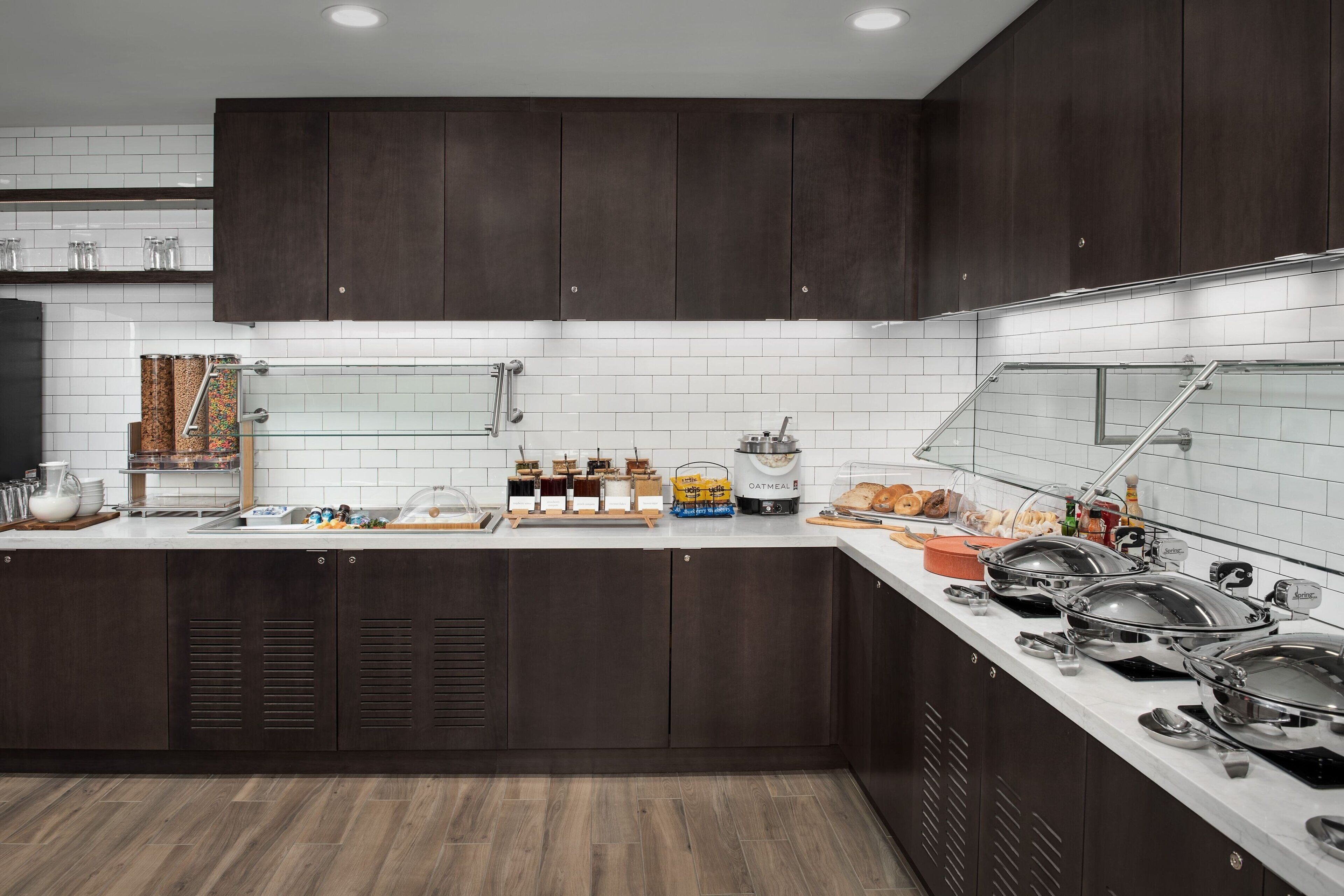 Una cocina moderna con armarios de madera oscura, salpicadero de azulejos blancos y diversas opciones para el desayuno sobre la encimera.