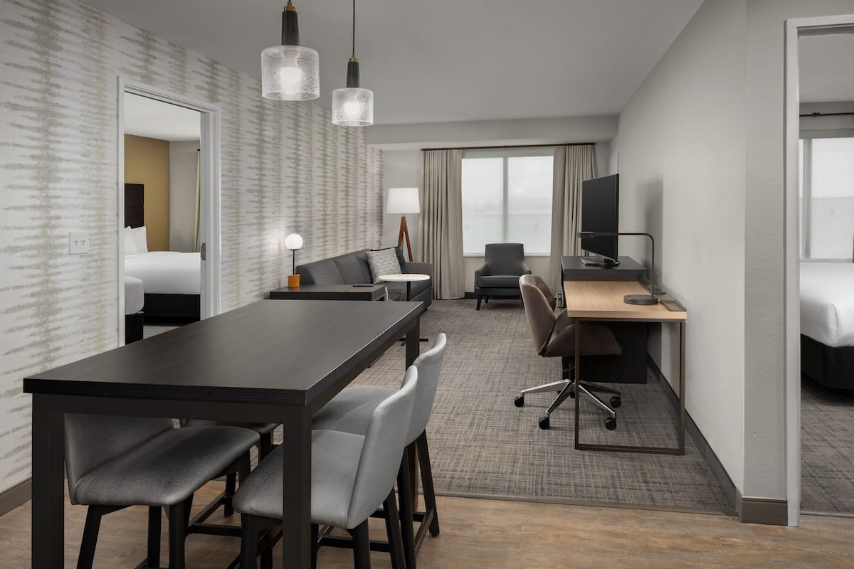 Ein Hotelzimmer mit dunklem Holzbüro, zwei grauen Stühlen, einem schwarzen Bürostuhl, einem Bett mit weißer Bettwäsche, einem Fernseher auf einem Standfuß und einem großen Fenster mit Vorhängen.