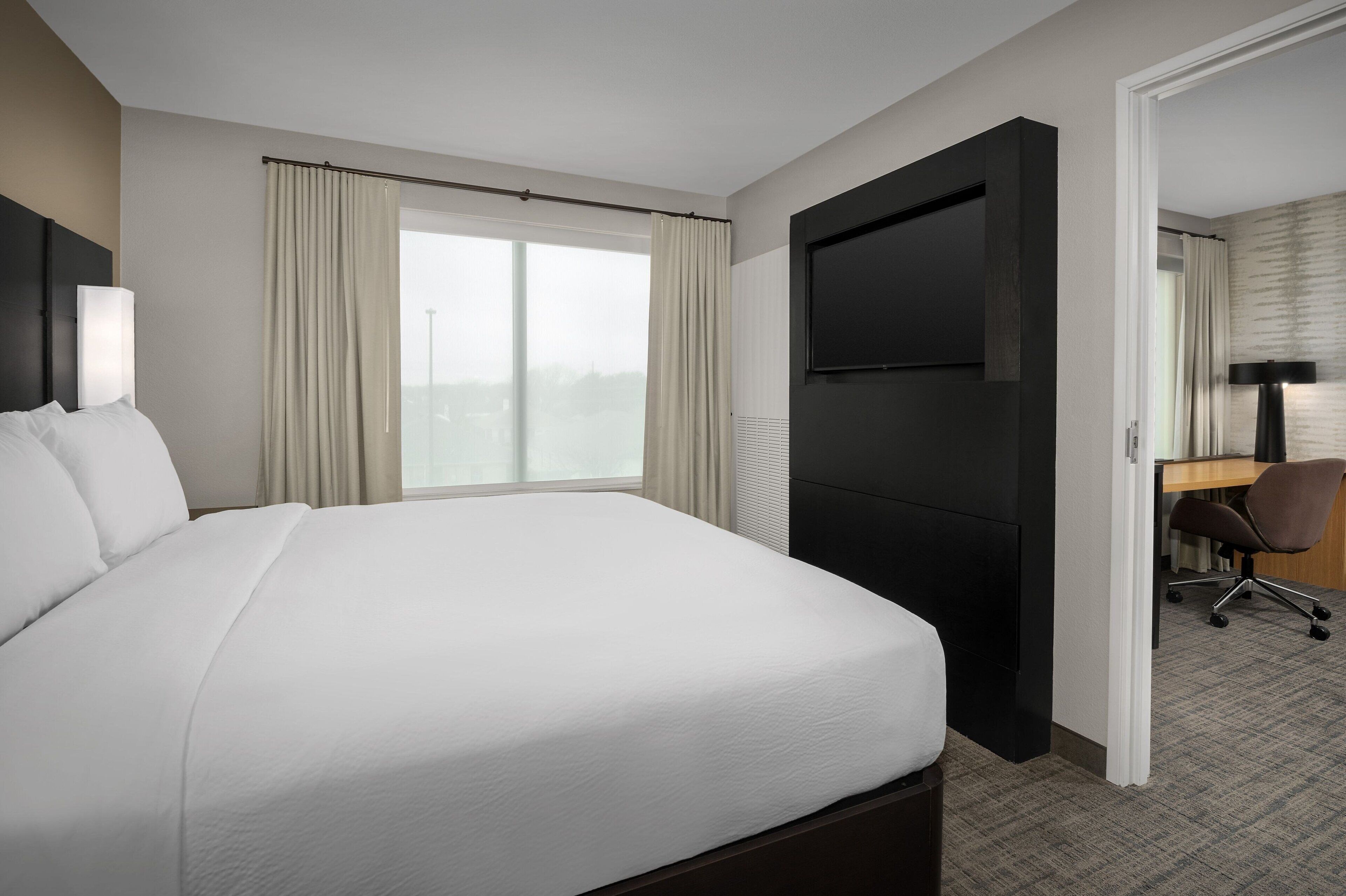 Habitación de hotel con una cama grande, un escritorio con silla, un televisor de pantalla plana y vistas a la ciudad desde la ventana.
