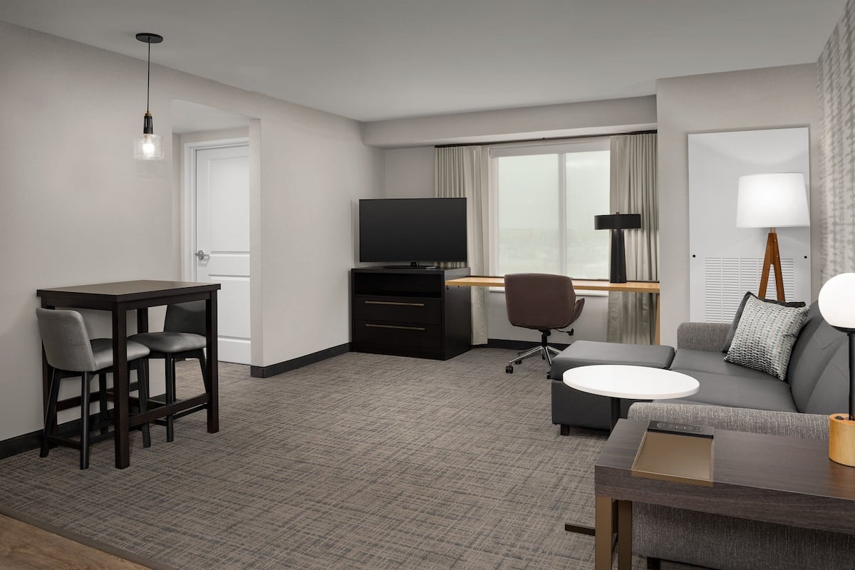 Ein modernes Hotelzimmer mit Flachbildfernseher, Schreibtisch mit Stuhl, einer Sitzecke und einem kleinen runden Tisch sowie einer Lampe.