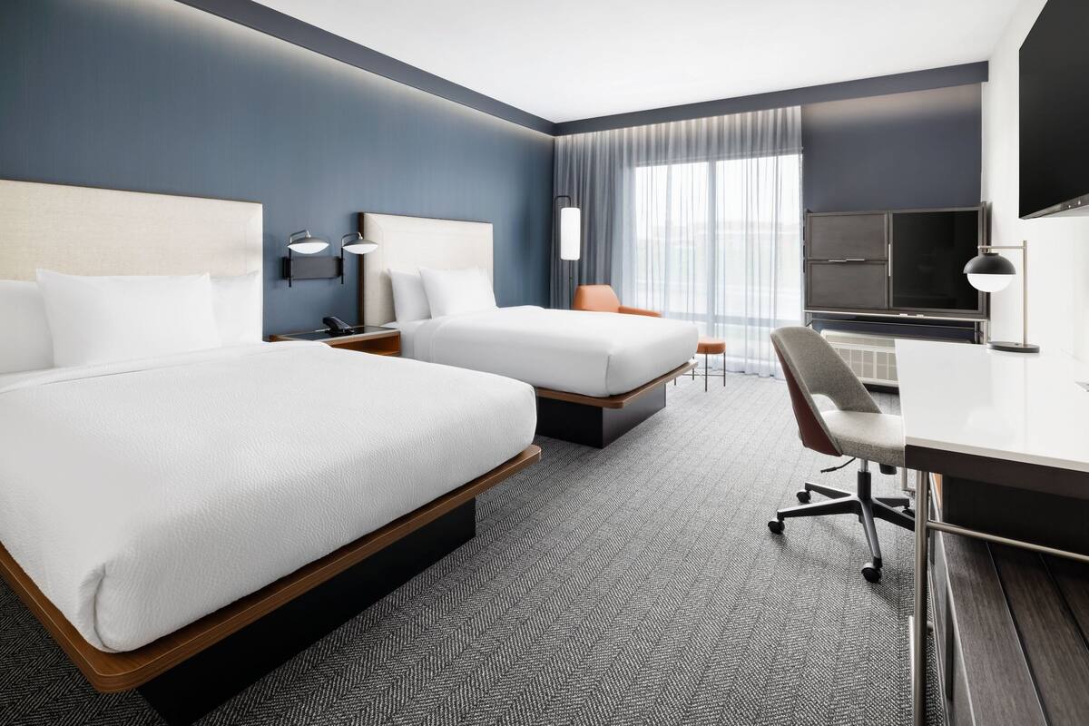 Ett hotellrum med två sängar, ett skrivbord, en stol, en TV och ett stort fönster med gardiner.