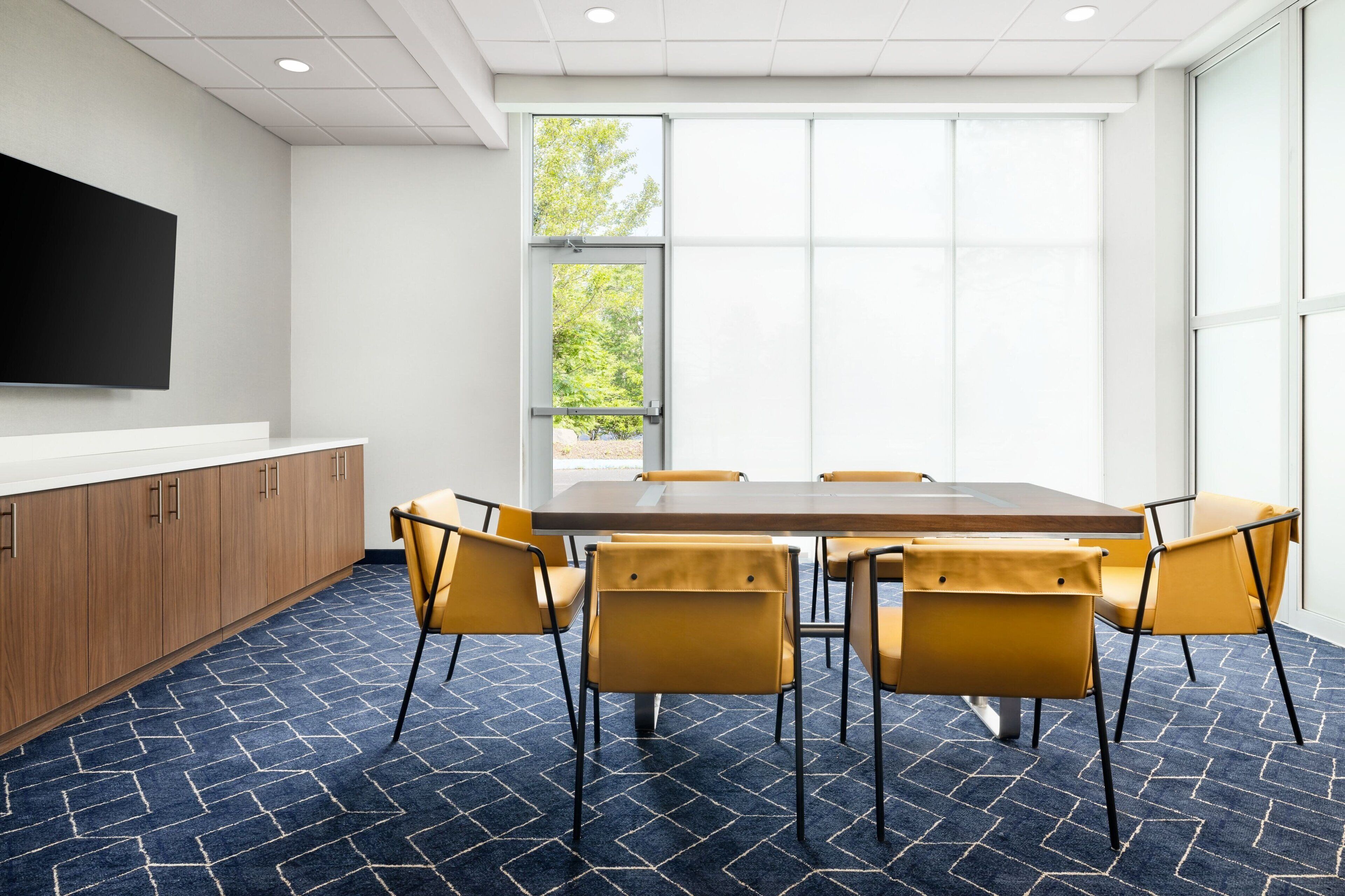 A conference room with a large table, six chairs, a flat-screen TV mounted on the wall, and large windows with a view of trees.