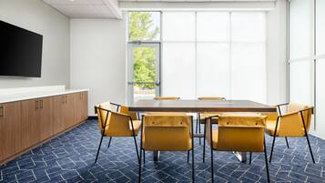 A conference room with a large table, six chairs, a flat-screen TV mounted on the wall, and large windows with a view of trees.