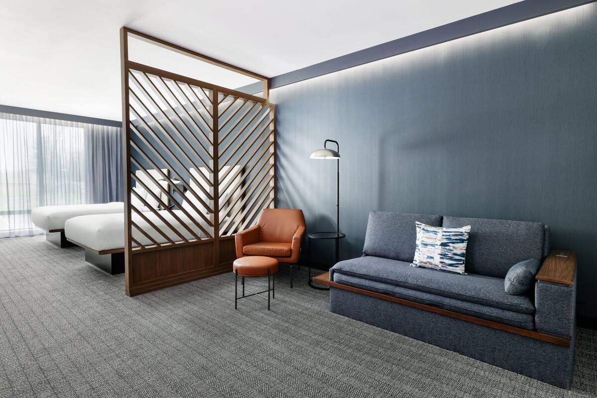 Ett modernt hotellrum med en grå soffa, en orange fåtölj, en vägg av trä och en golvlampa.