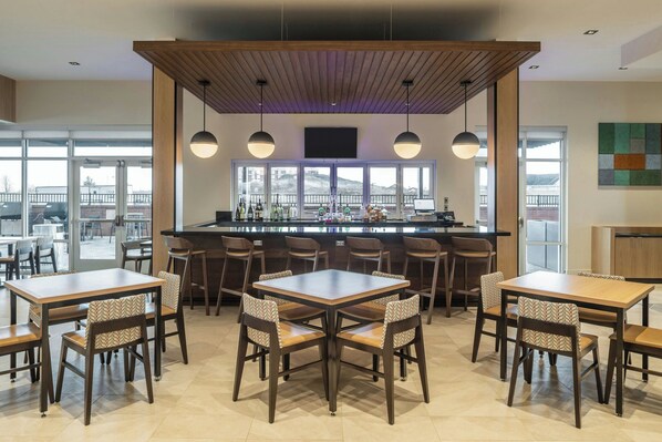 A modern dining area with wooden tables and chairs, a bar counter, and large windows offering a view of the outdoors.