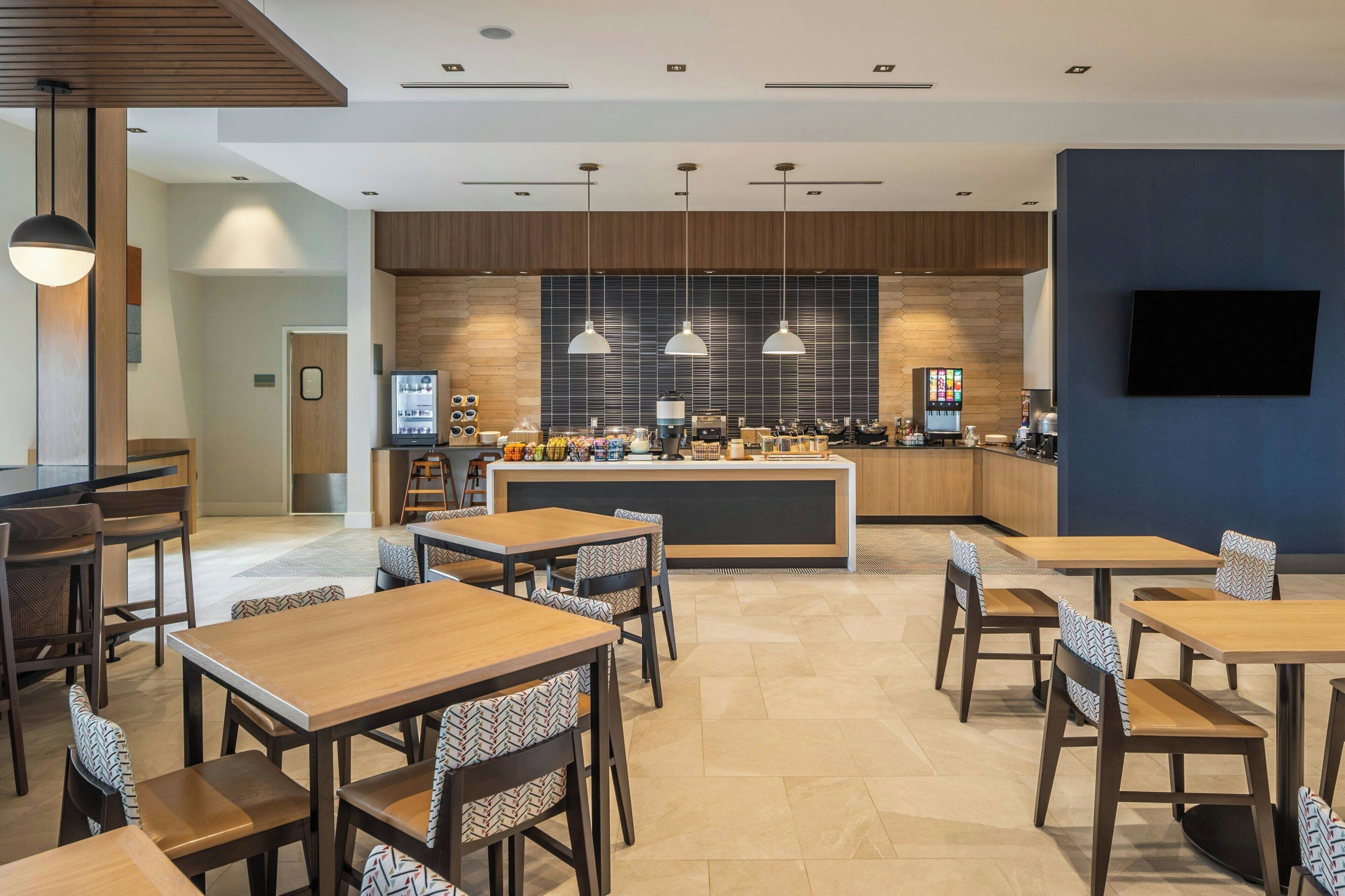 A modern restaurant with wooden tables and chairs, a central bar area, and pendant lighting.
