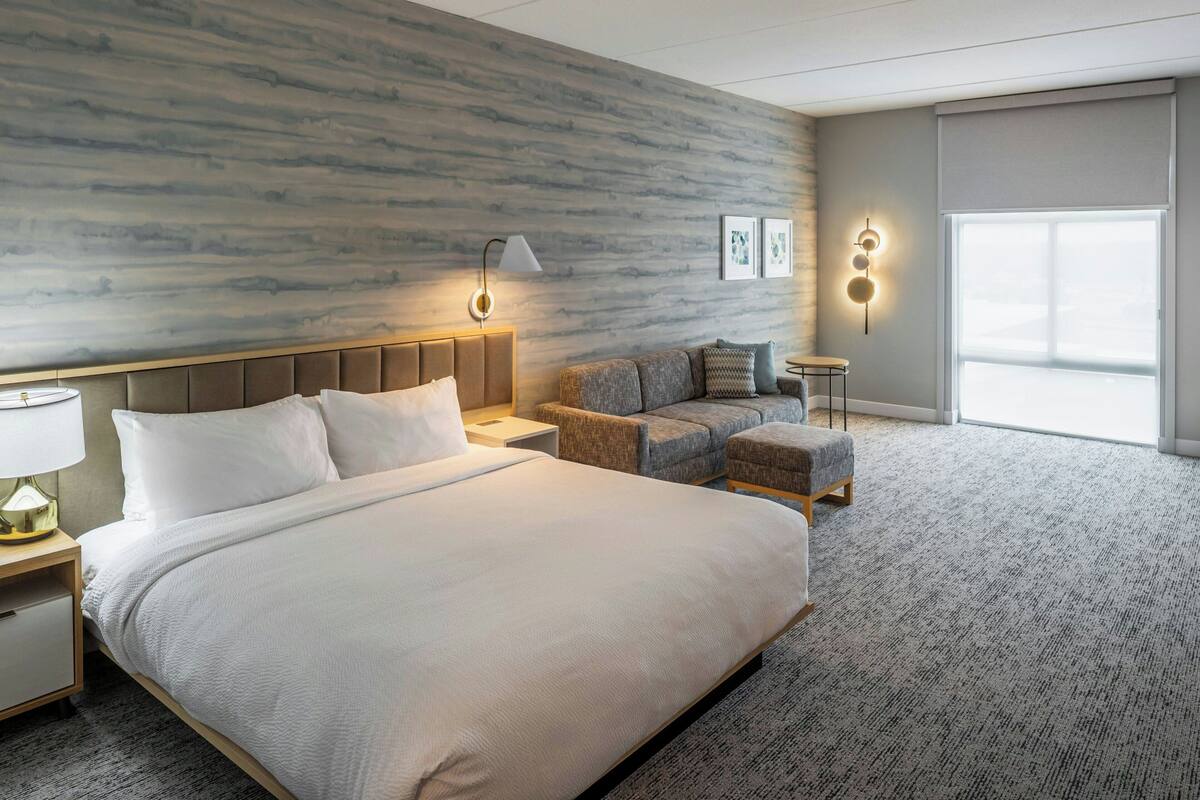A modern hotel room with a large bed, a sofa, a small table, and a nightstand.