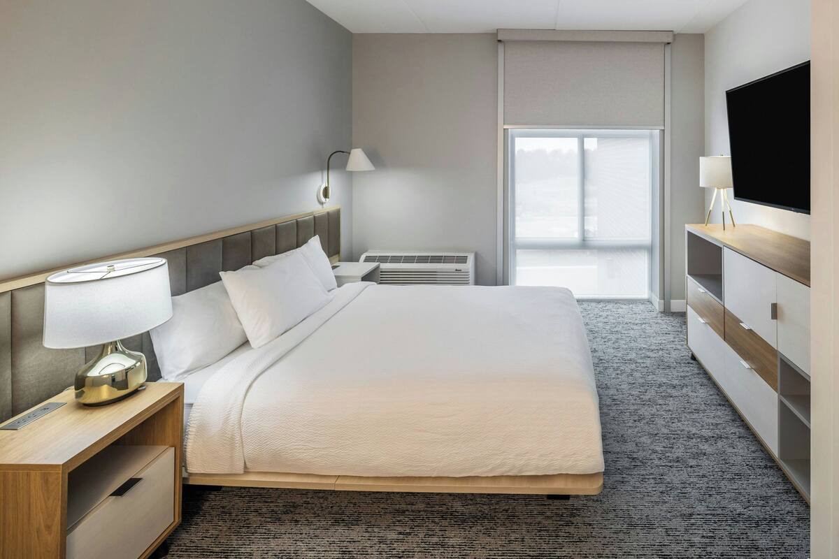A hotel room with a large bed, a bedside table with a lamp, a TV mounted on the wall, and a window with a view.