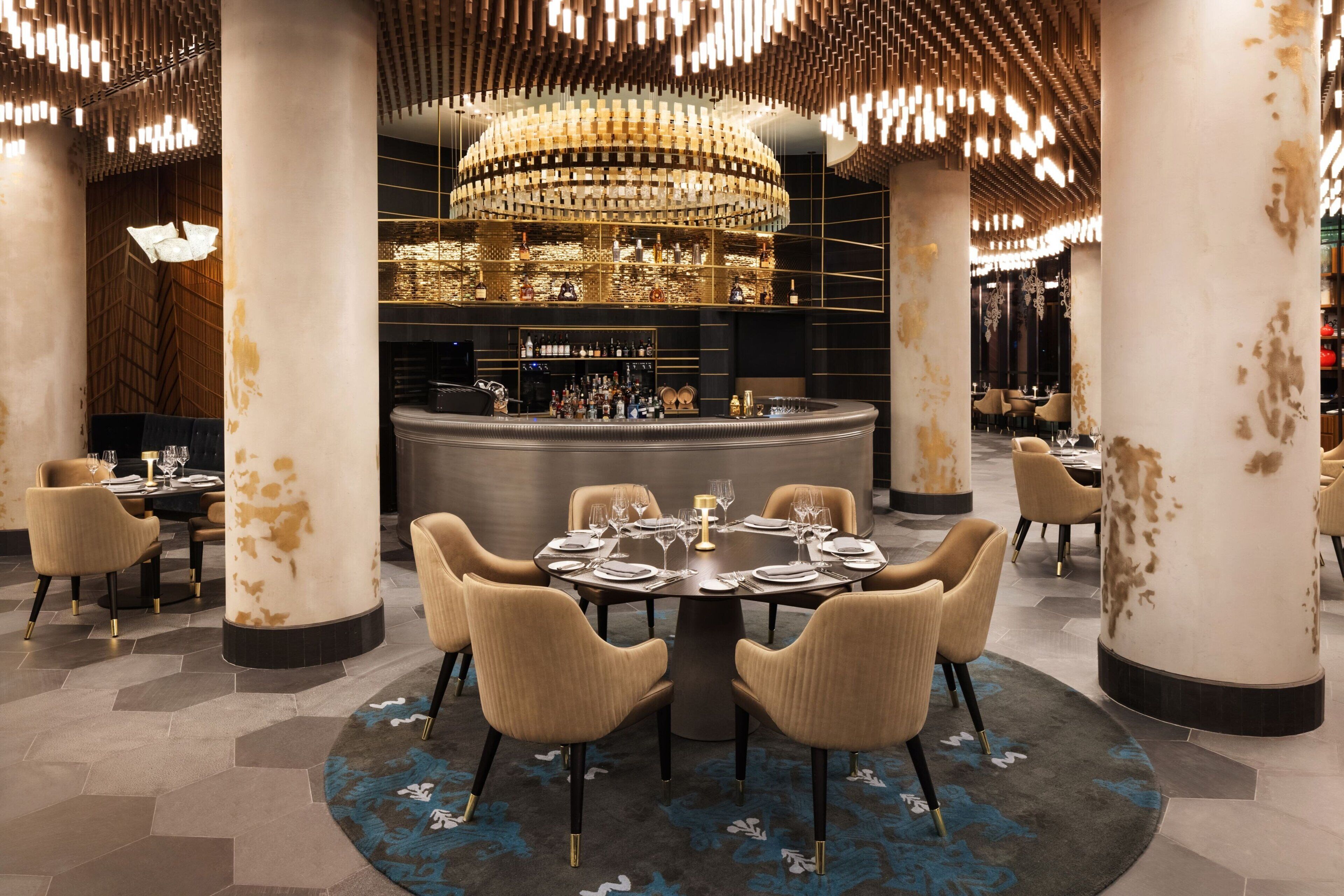 Un restaurante moderno con mesas redondas, sillas color beige y una zona de bar central.