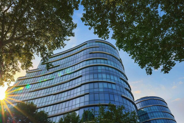 A modern curved glass building with multiple floors, surrounded by trees.
