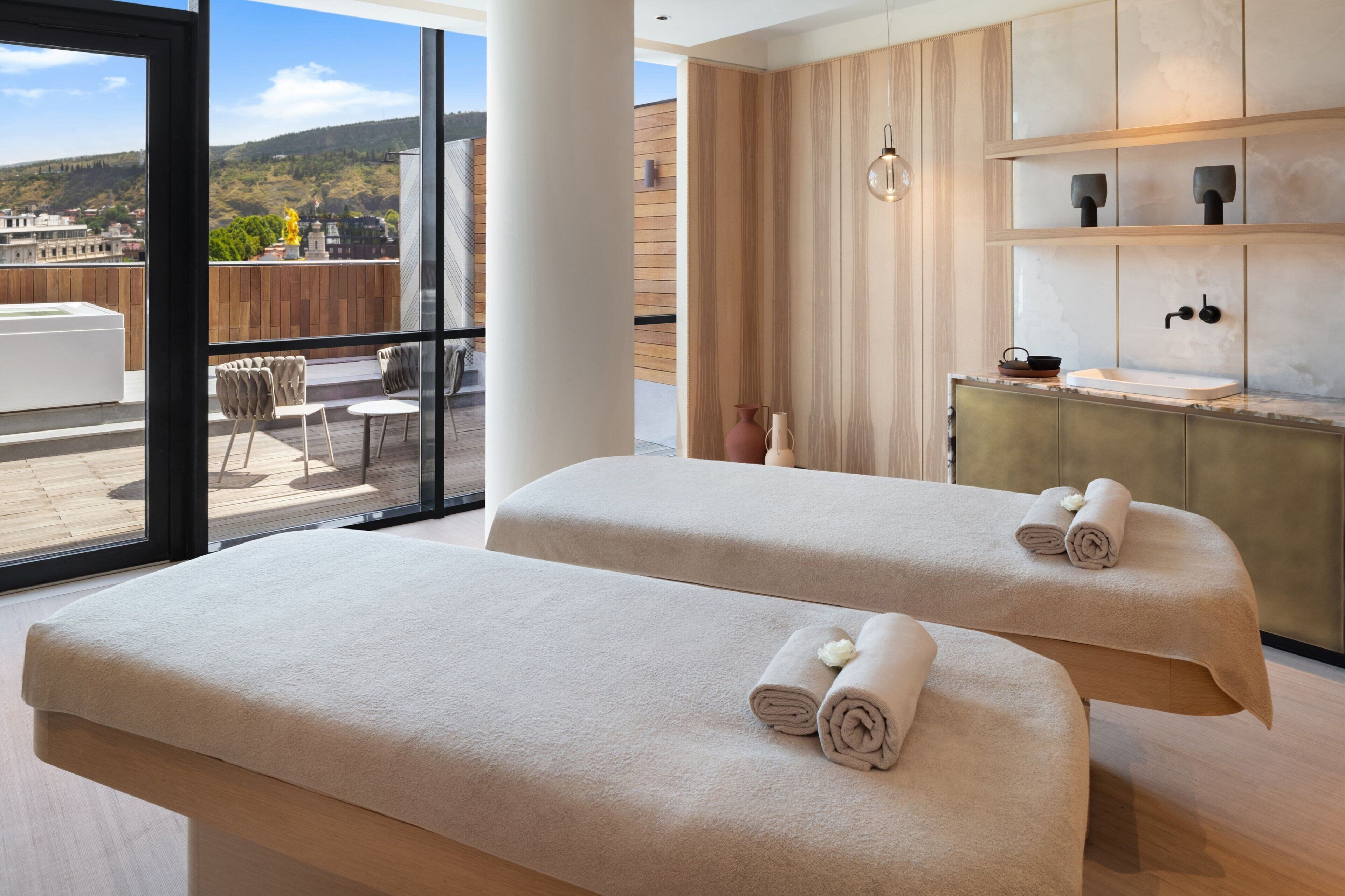 Un moderno spa con dos camillas de masaje, una pared de madera, un amplio ventanal con vista a la ciudad y una pequeña zona de lavamanos.