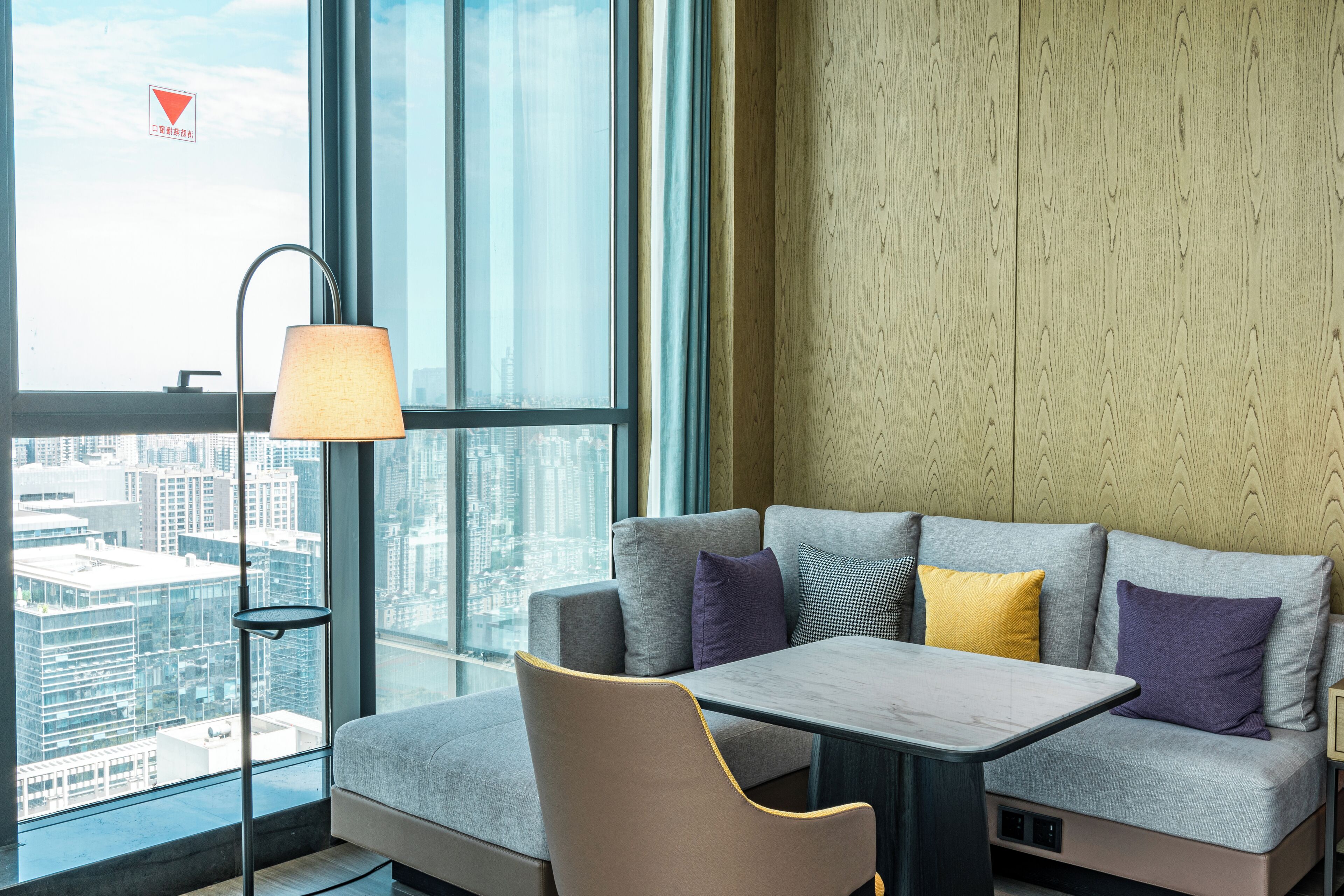 A modern hotel room with a large window offering a city view, a sofa with multiple cushions, a small table, and a floor lamp.