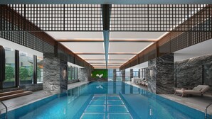 An indoor swimming pool with a clear view of the pool area, a seating area with lounge chairs, and a view of the outside through large windows.