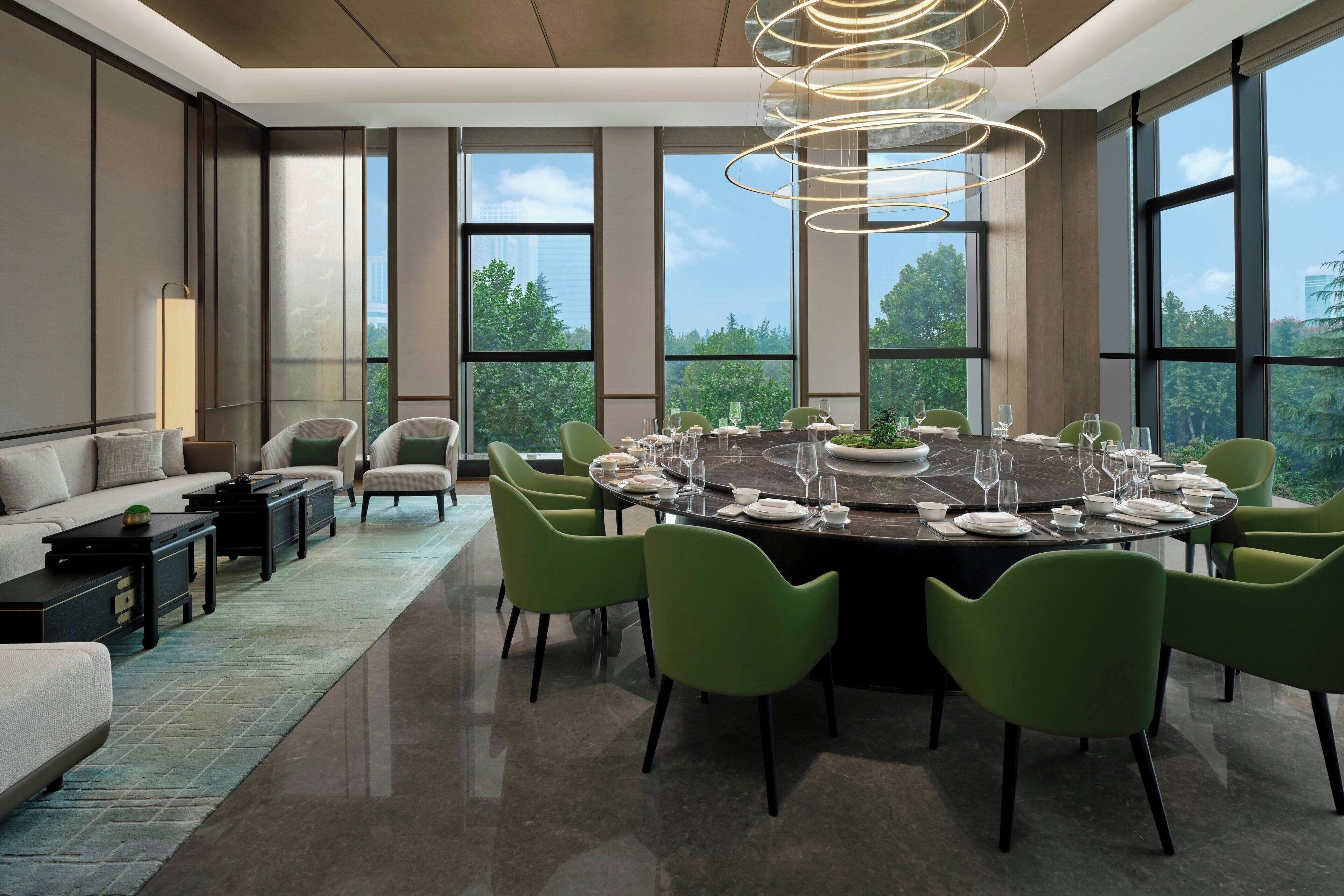 A modern dining area with a round table set for six, surrounded by green chairs, large windows offering a view of trees and buildings, and a contemporary light fixture above.