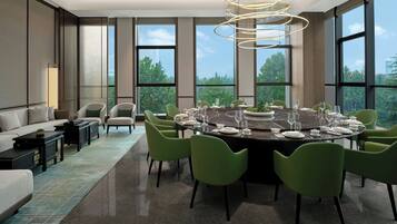 A modern dining area with a round table set for six, surrounded by green chairs, large windows offering a view of trees and buildings, and a contemporary light fixture above.