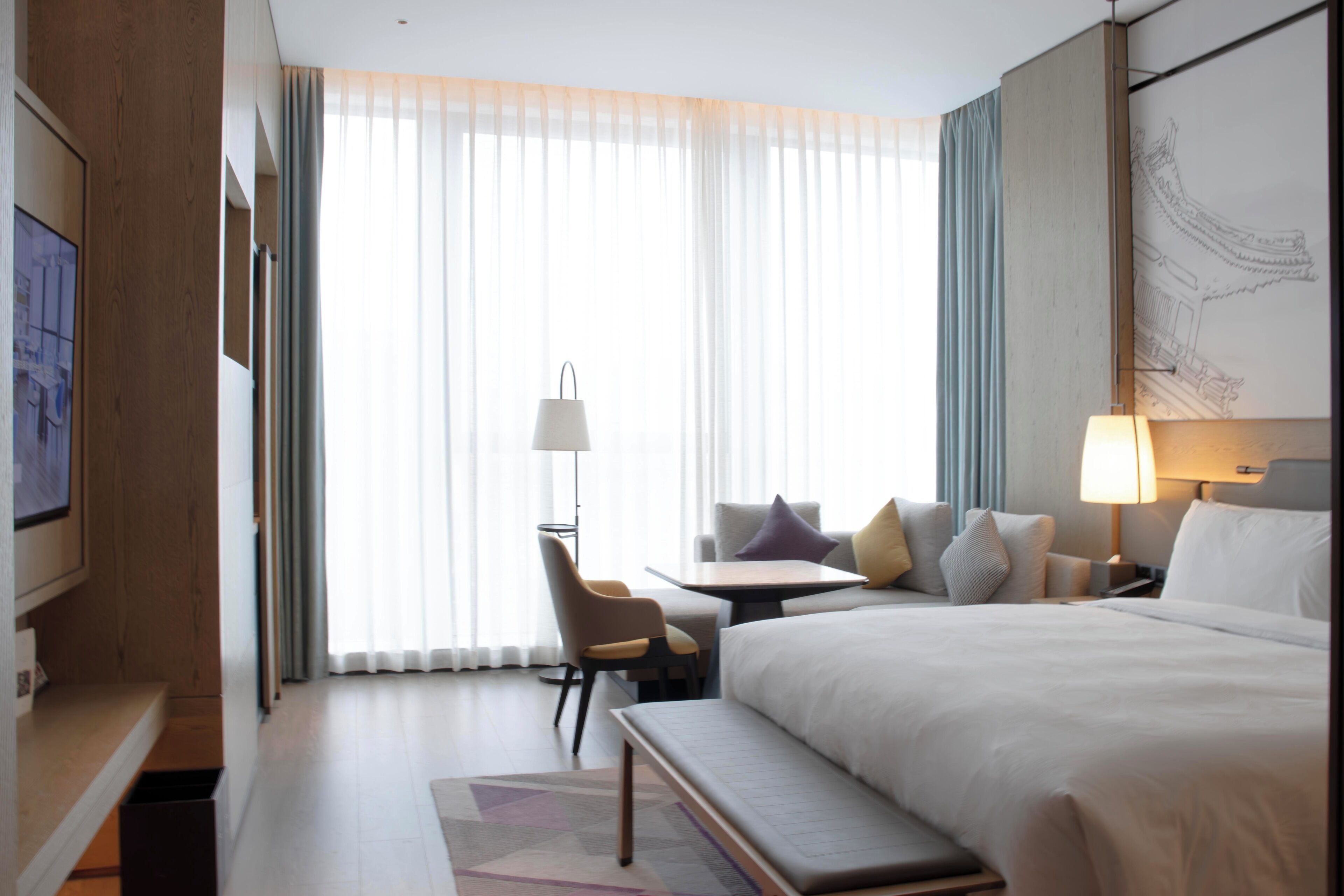 Una habitación de hotel con una cama grande, un escritorio, una silla y una ventana con cortinas translúcidas.