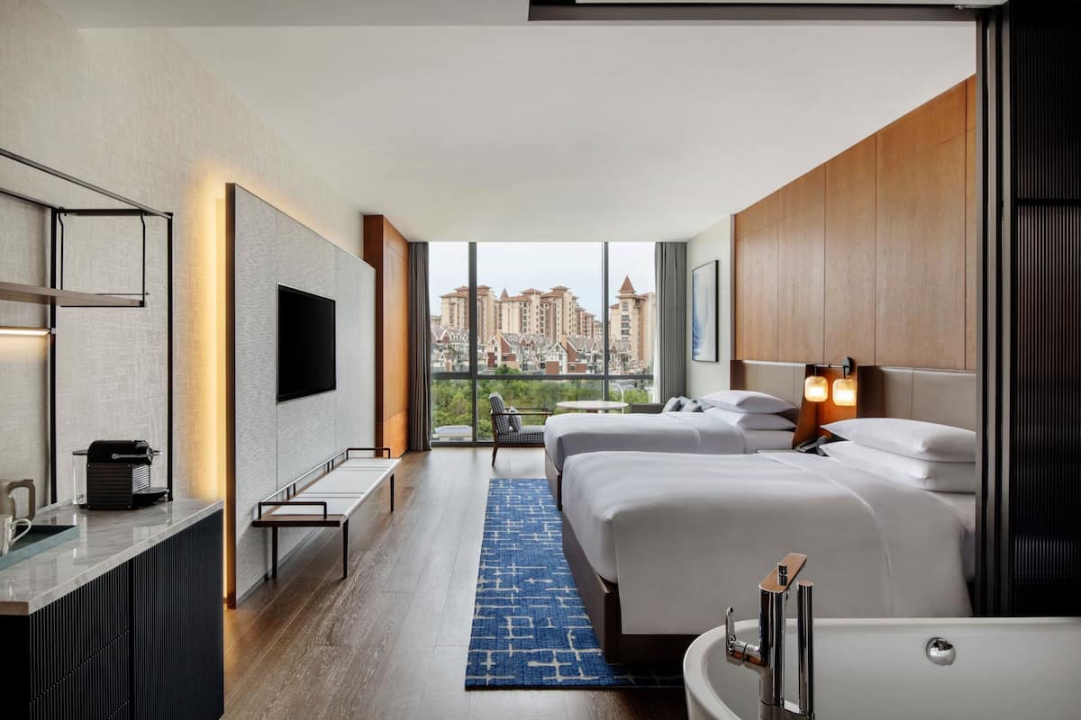A modern hotel room with a large window, two beds, a desk, and a bathroom.