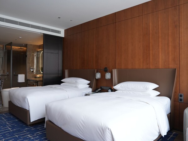 Habitación de hotel con dos camas, un panel de pared de madera, un baño con espejo y una alfombra azul estampada.