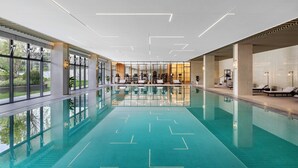 Una piscina interior moderna con paredes de vidrio transparente, un diseño geométrico en el suelo y una zona central con sillones de descanso.