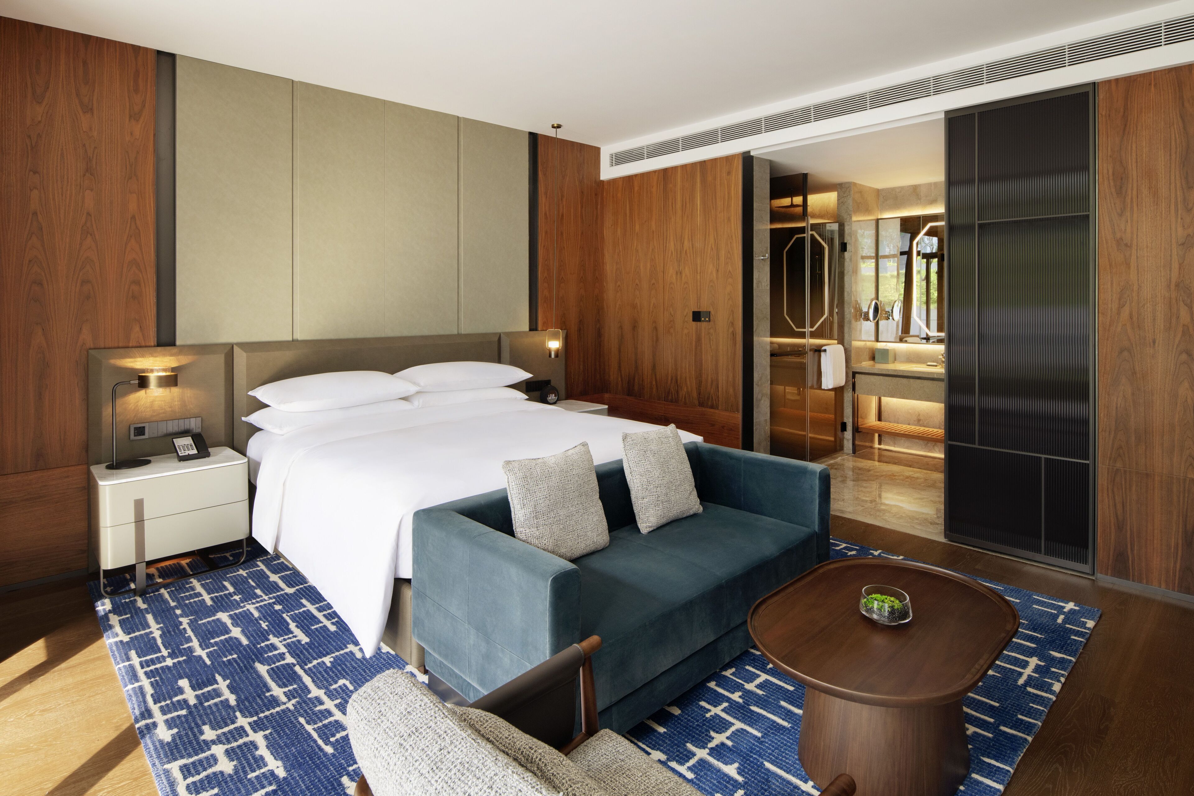 Habitación de hotel moderna con una cama grande, un sofá, una mesita y un baño visible a través de una puerta abierta.