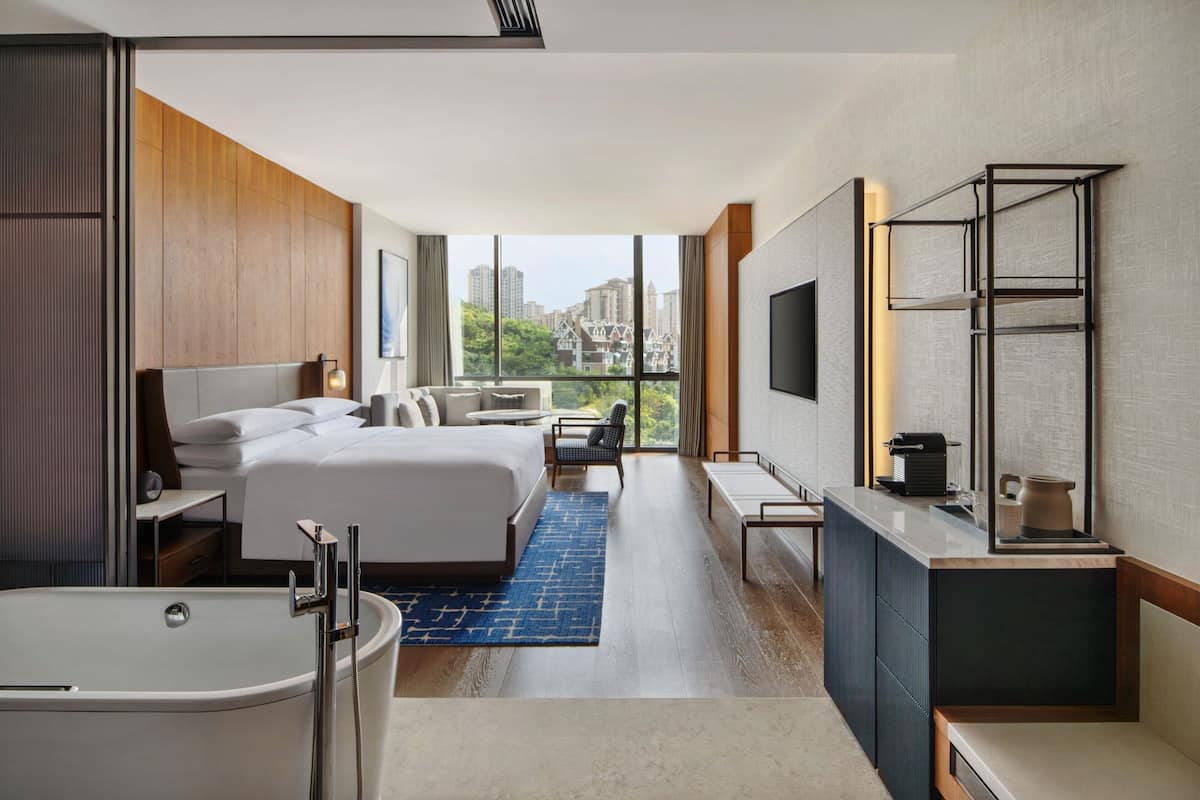 A modern hotel room with a large bed, a separate seating area, a bathroom with a bathtub, and a city view.
