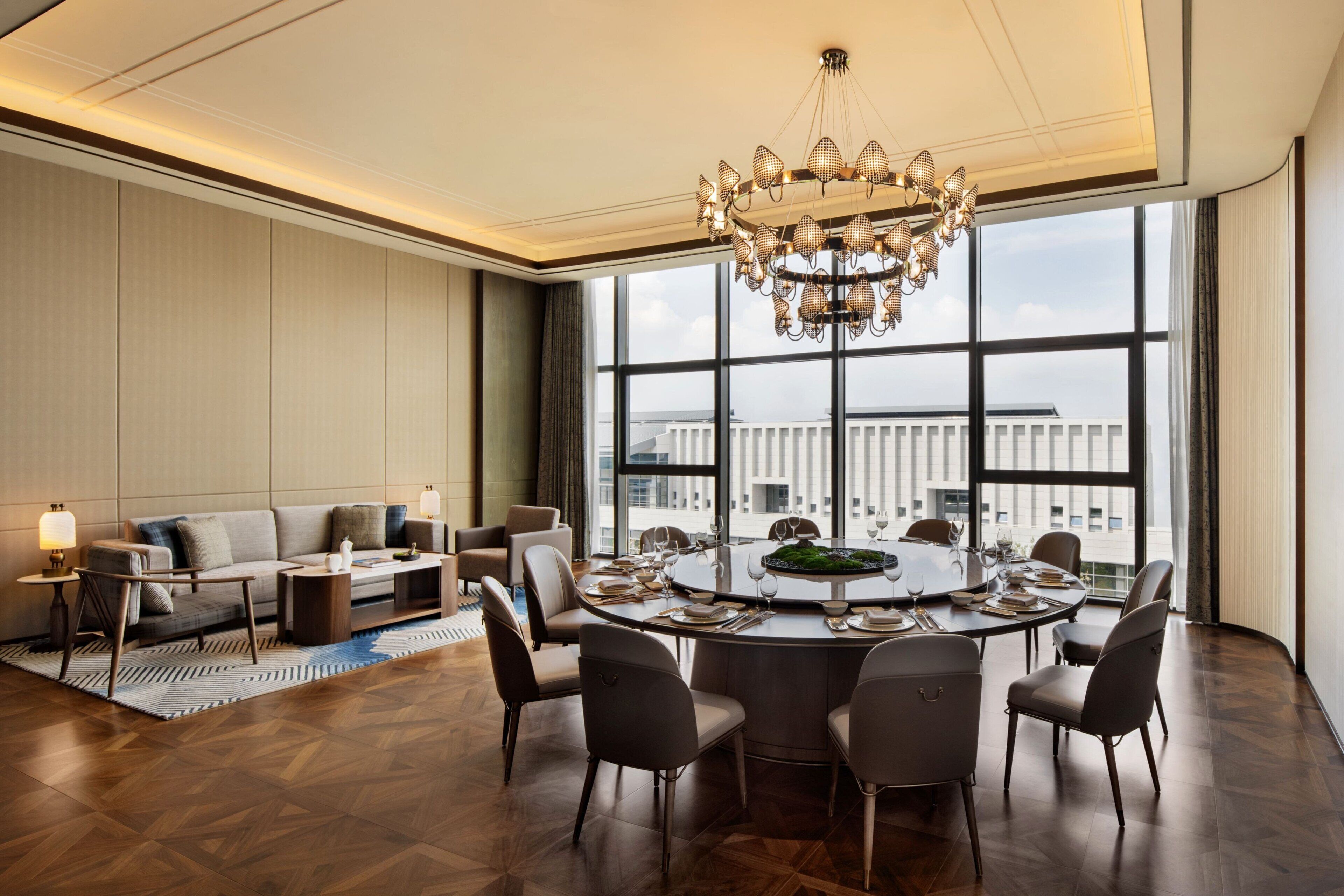 A modern hotel room with a round dining table, comfortable seating, a chandelier, and large windows offering a city view.
