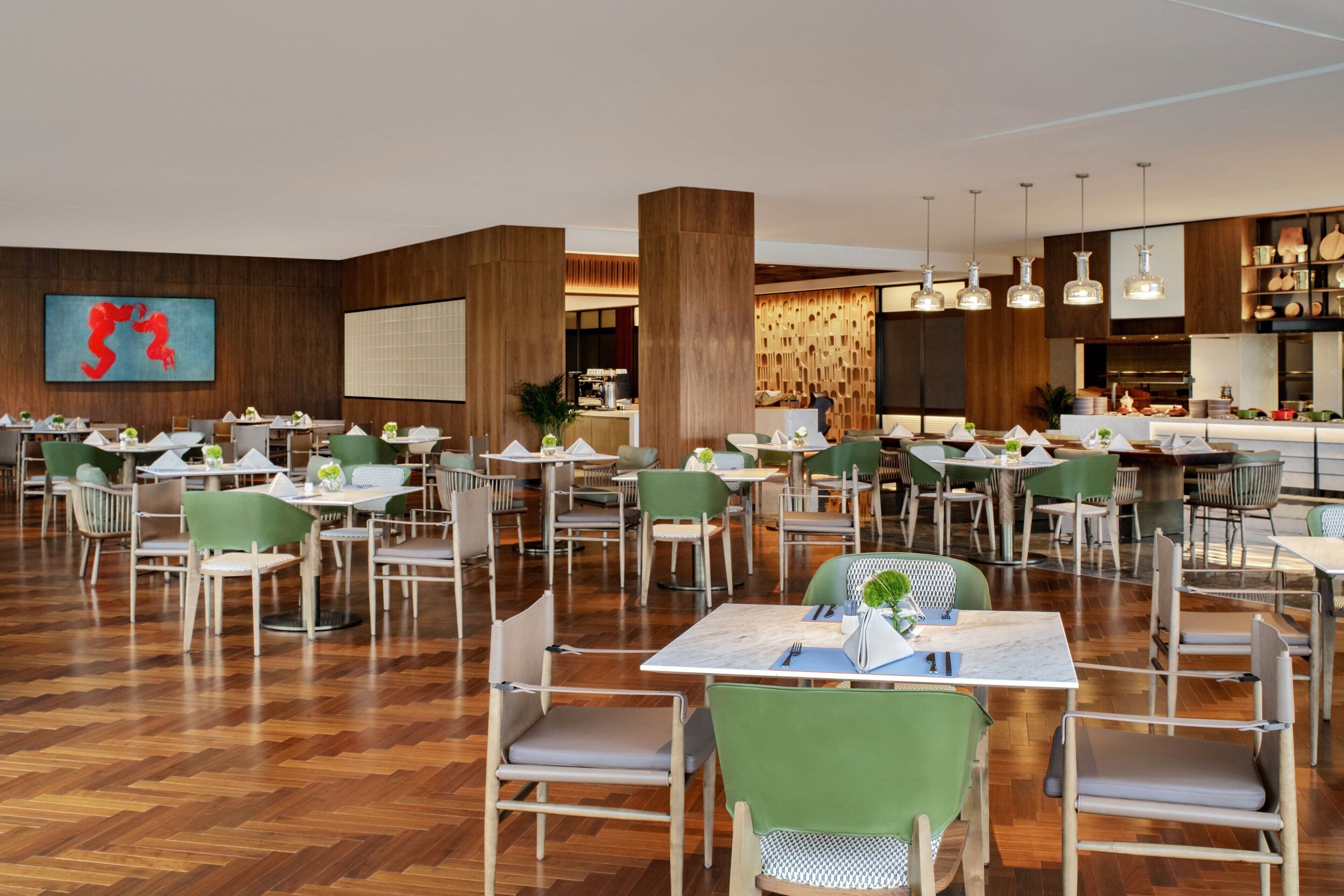 A spacious dining area with multiple tables and chairs, wooden flooring, and a modern kitchen in the background.