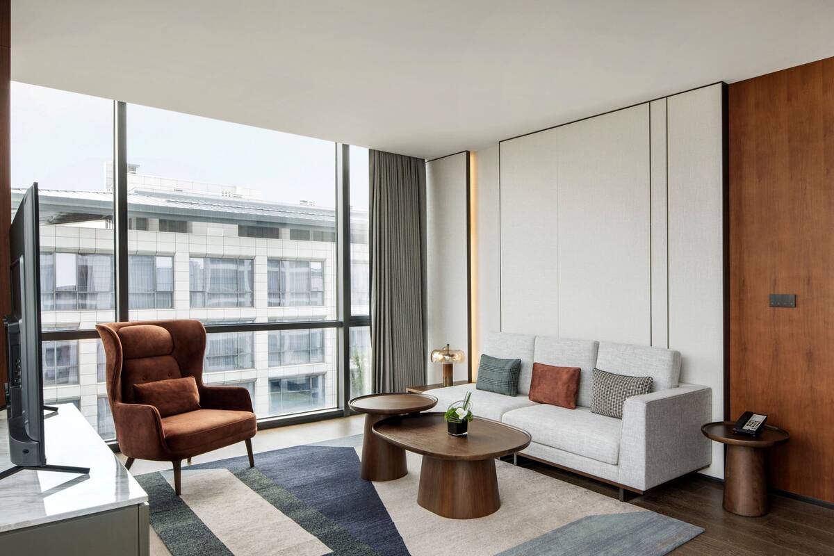 A modern hotel room with a large window, a sofa, a chair, a coffee table, and a side table with a phone.