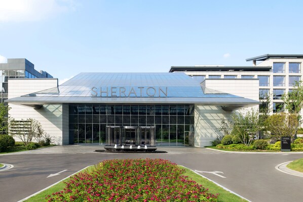 A Sheraton hotel entrance with a glass facade, a fountain in front, and a landscaped garden area. - Sheraton Xuzhou (Xuzhou)