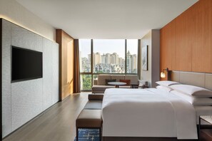 A modern hotel room with a large bed, a flat-screen TV mounted on the wall, a seating area with a sofa and chairs, and a view of a cityscape through large windows. - Sheraton Xuzhou (Xuzhou)