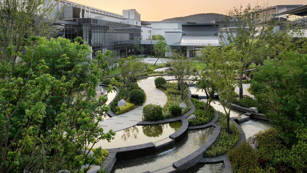 Un moderno complejo de edificios con un jardín ajardinado que incluye una fuente sinuosa y varias especies de árboles.
