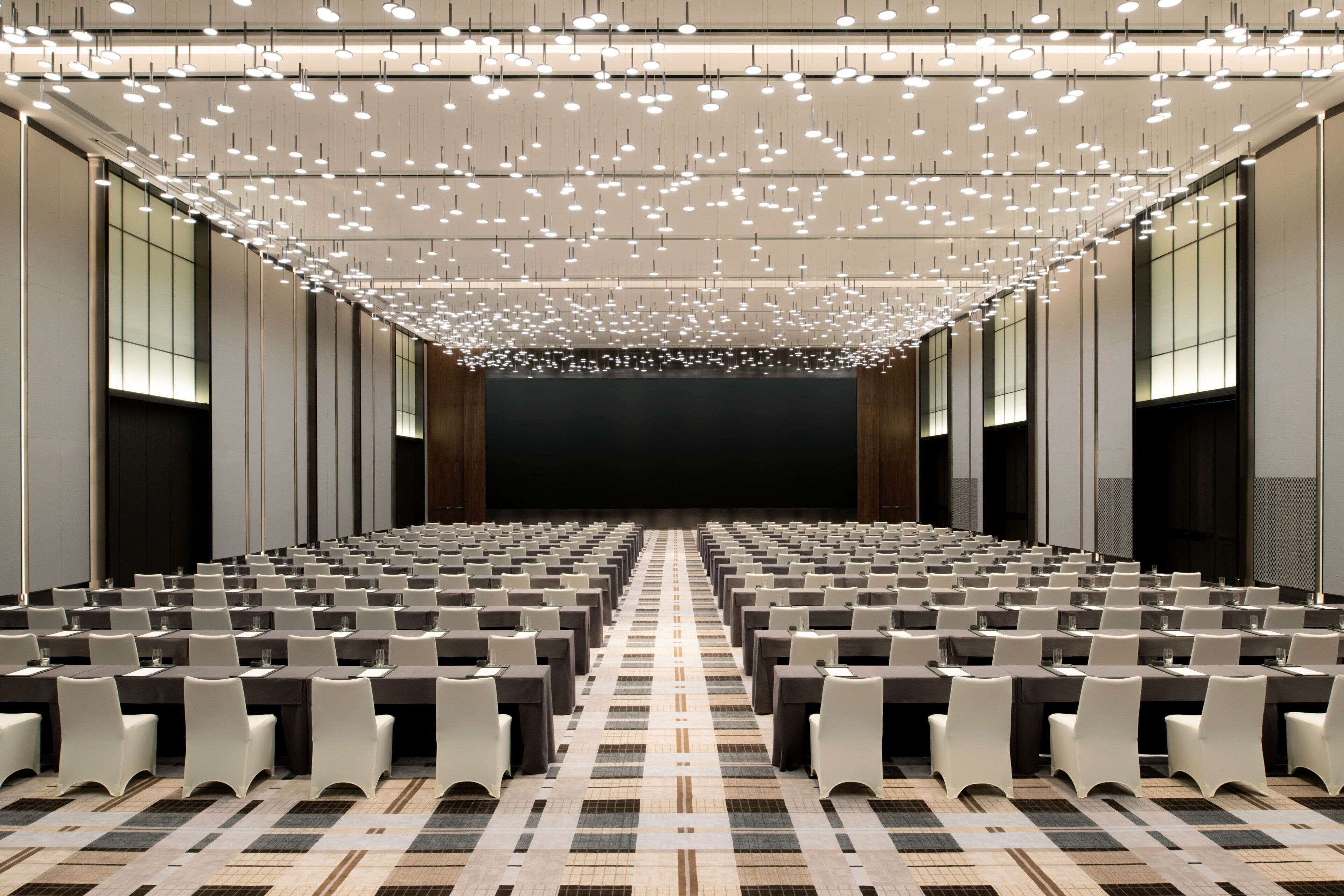 A conference room with rows of tables and chairs, a patterned carpet, and a ceiling with numerous hanging lights.
