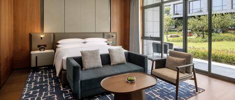 A modern hotel room with a large bed, a sofa, a coffee table, and a view of the garden through large windows.