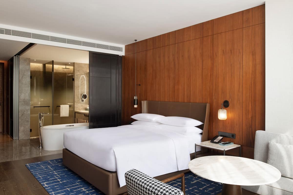 A modern hotel room with a large bed, a bedside table with a phone, a round table, a sofa, and a bathroom with a bathtub and shower.