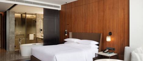 A modern hotel room with a large bed, a bedside table with a phone, a round table, a sofa, and a bathroom with a bathtub and shower.