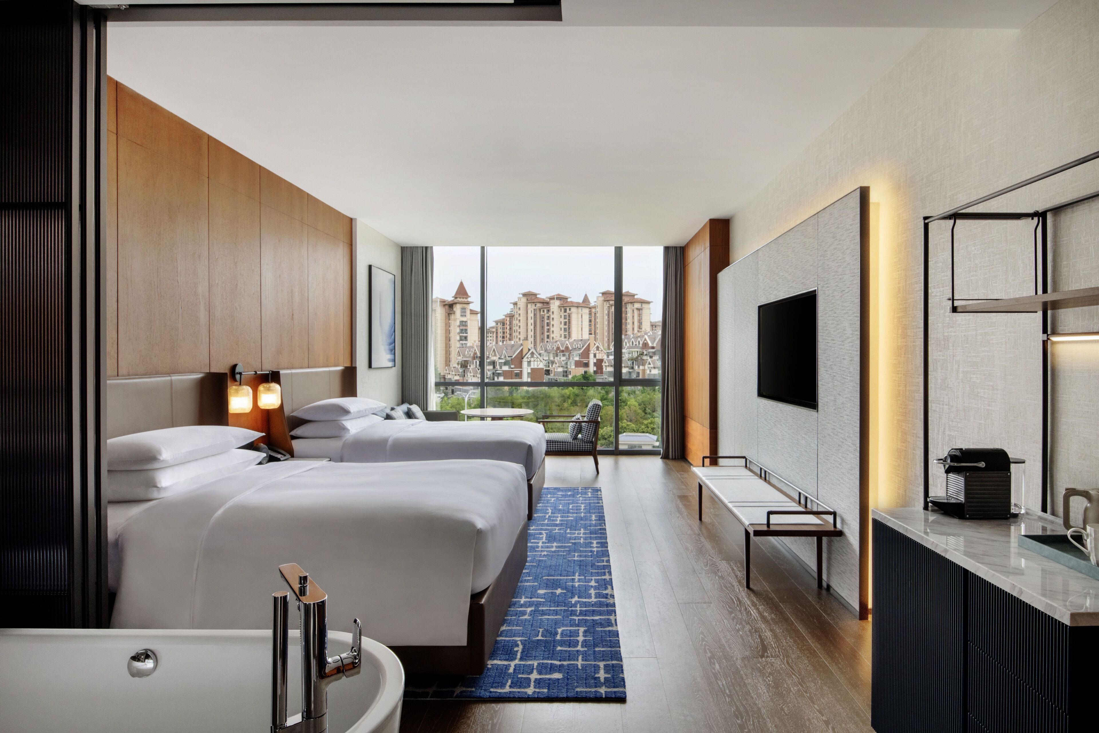 A hotel room with two beds, a large window with a city view, a flat-screen TV, and a small kitchenette.