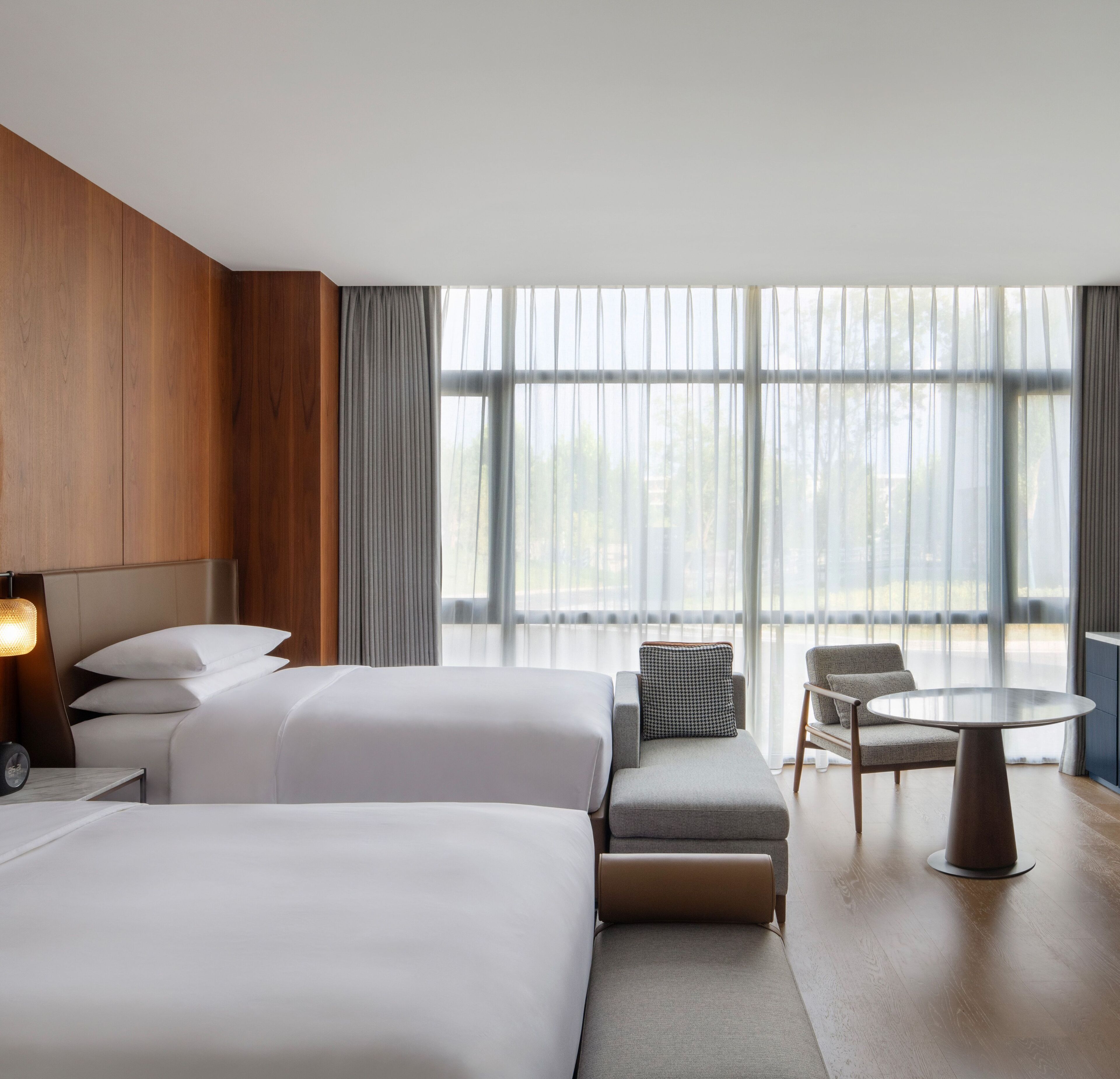 A modern hotel room with two large beds, a sofa, a small table, and a view of the outdoors through large windows.