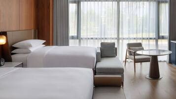 A modern hotel room with two large beds, a sofa, a small table, and a view of the outdoors through large windows.