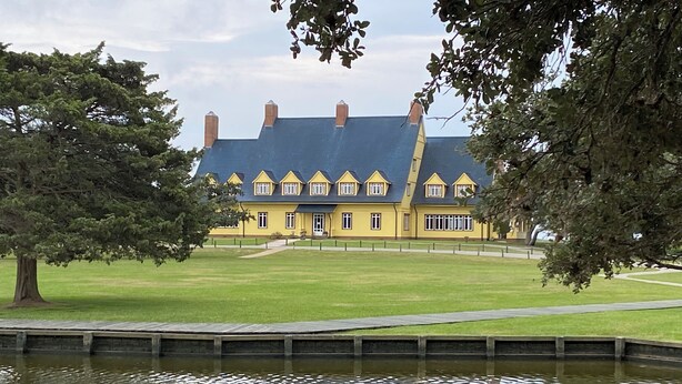 A large yellow building with a blue roof, surrounded by a grassy area and a body of water. - EXCEPTIONAL LOCATION - LUXURY VILLA STEPS AWAY FROM THE BEACH AND POOL (Corolla)