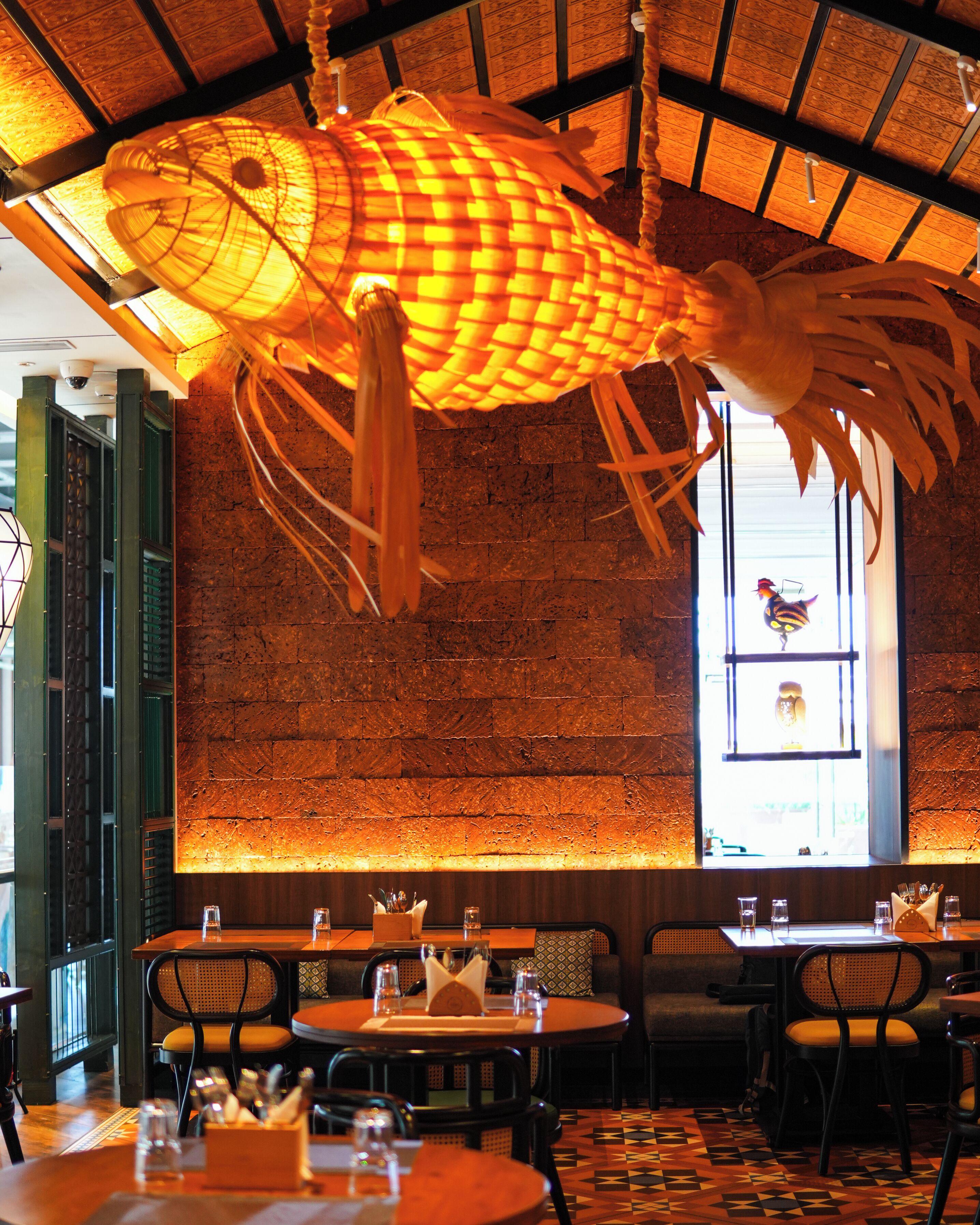 Interior de restaurante con una gran escultura de pez colgante, paredes de ladrillo y suelo con diseño. Mesas y sillas dispuestas para comer.