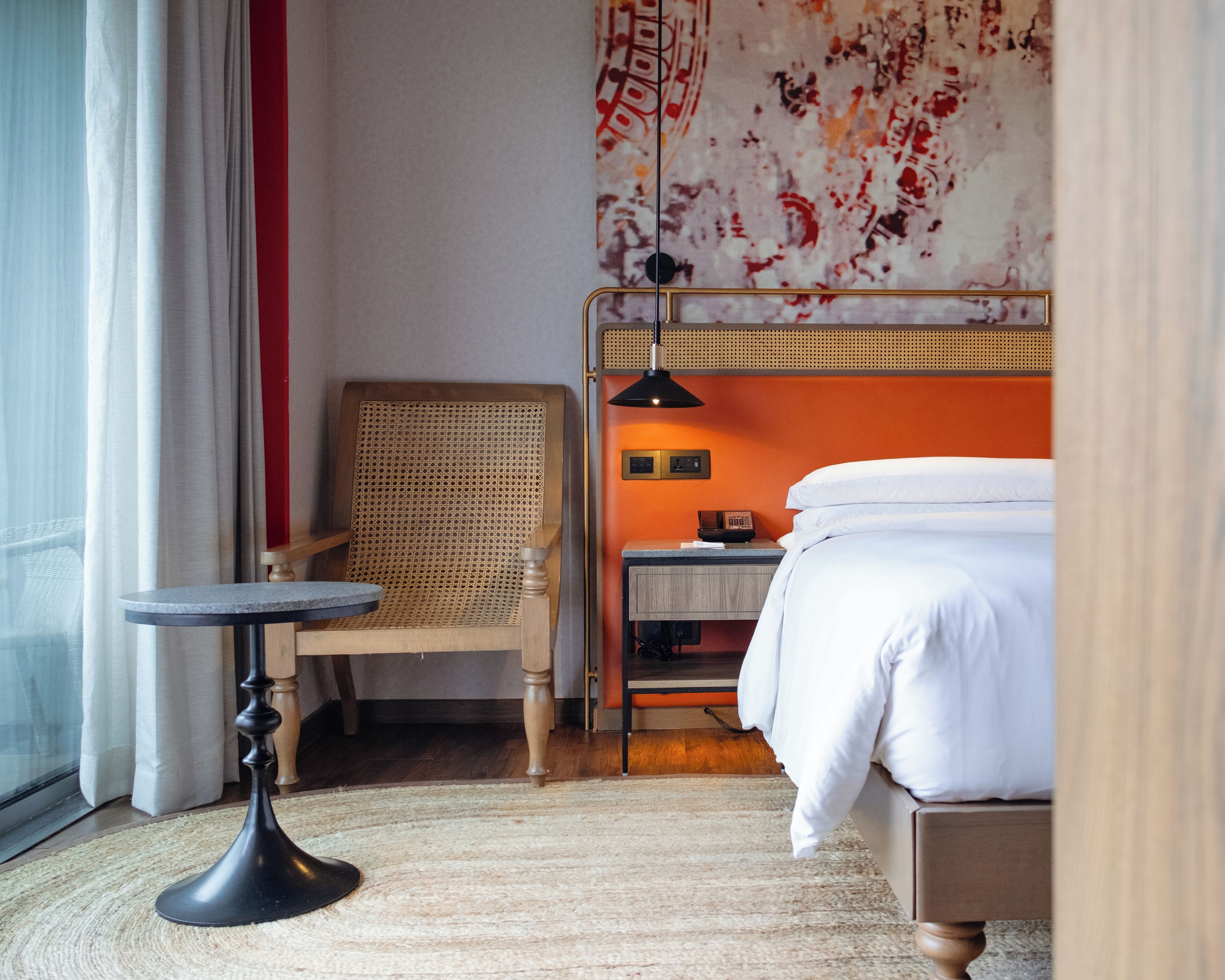 A hotel room with a bed, a chair, a small table, a lamp, and a wall with a decorative pattern.