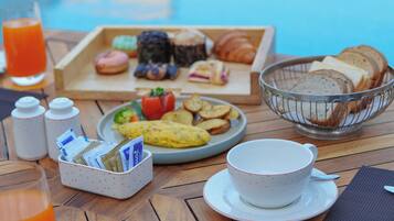Desayuno junto a una piscina, con diversos pasteles, pan y un vaso de zumo de naranja.