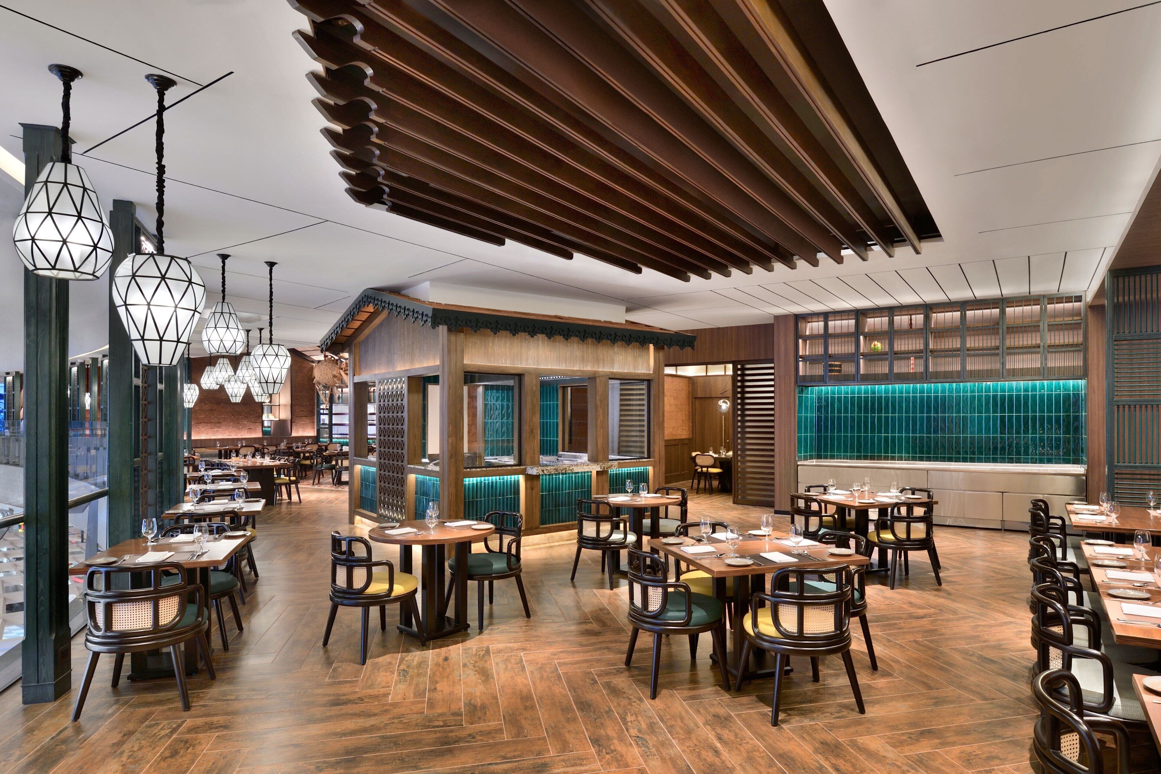 A spacious restaurant with wooden flooring, multiple dining tables, and hanging geometric light fixtures.