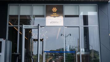 Une entrée moderne avec des portes en verre, une enseigne indiquant « The Artististe Tribute Portfolio Marriott Bonvoy », et le reflet de l’environnement dans le verre.