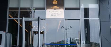 Entrada moderna de un edificio con puertas de vidrio, un cartel que dice "The Artististe Tribute Portfolio Marriott Bonvoy" y el reflejo de los alrededores en el vidrio.