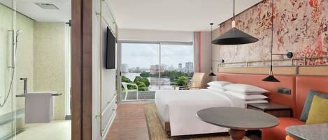 A modern hotel room with a large bed, a small table, a sofa, a TV, and a balcony with a city view.