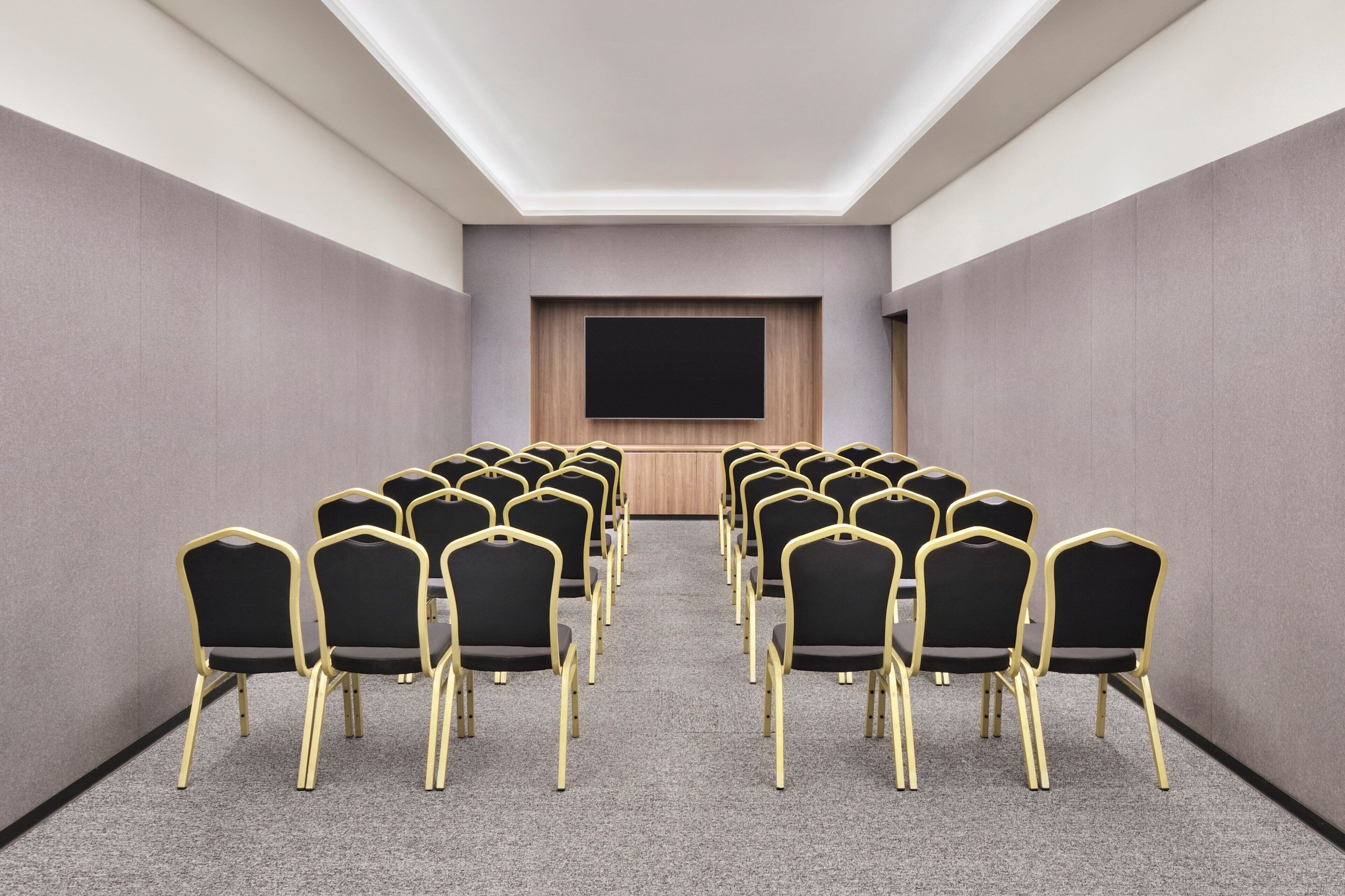 Un salón de conferencias con filas de sillas negras con detalles dorados, un televisor de pantalla plana grande montado en una pared de paneles de madera, y piso alfombrado.