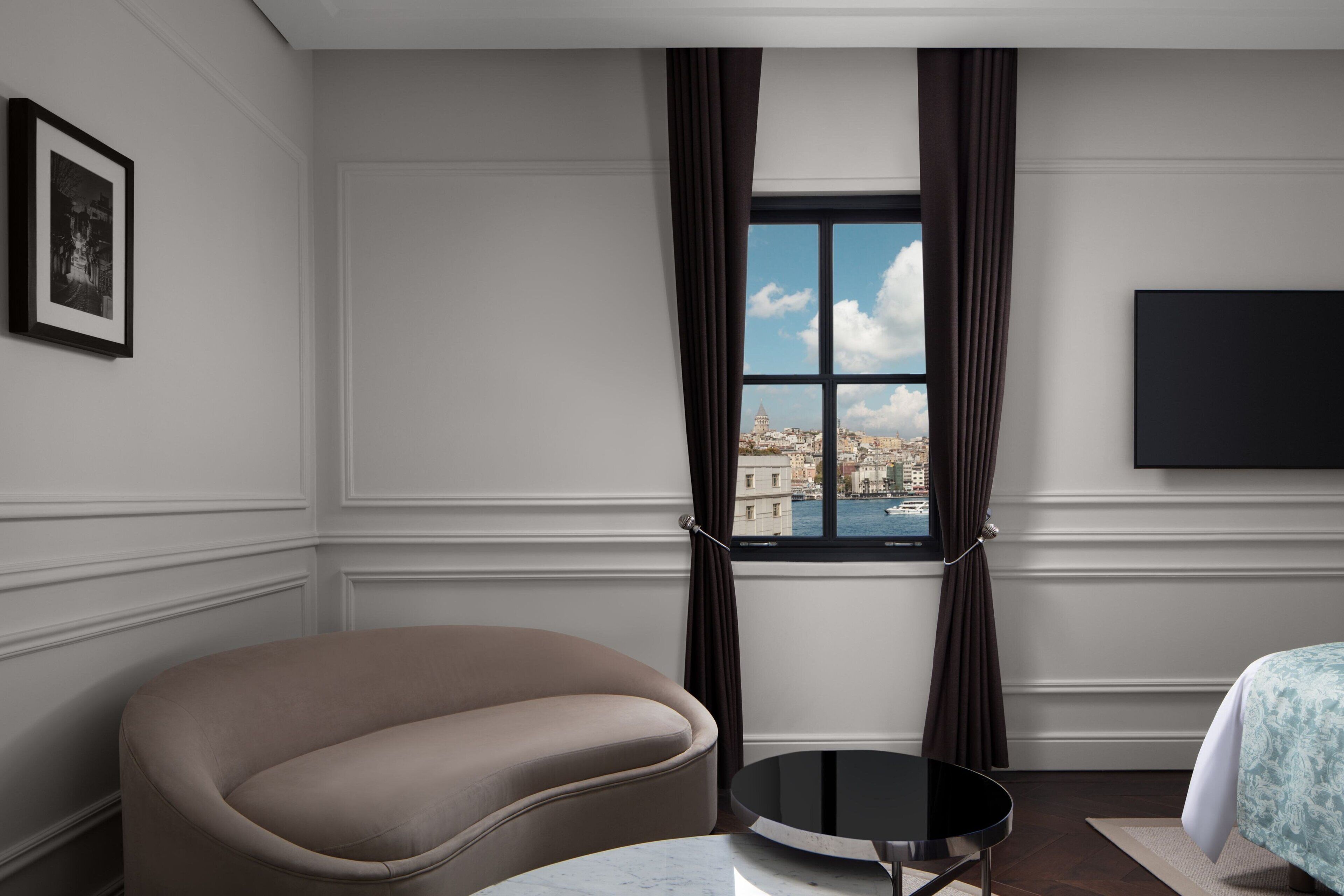 A modern hotel room with a curved sofa, a round table, a large window with curtains, a flat-screen TV, and a framed picture on the wall.